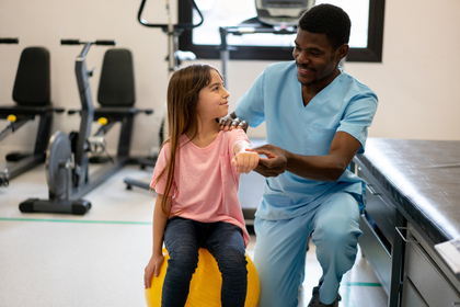 beautiful little girl looking at her therapist while he helps hold her arm and she keeps balance on a fitness ball